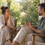Two adults listening and talking on a sunlit veranda surrounded by green foliage.